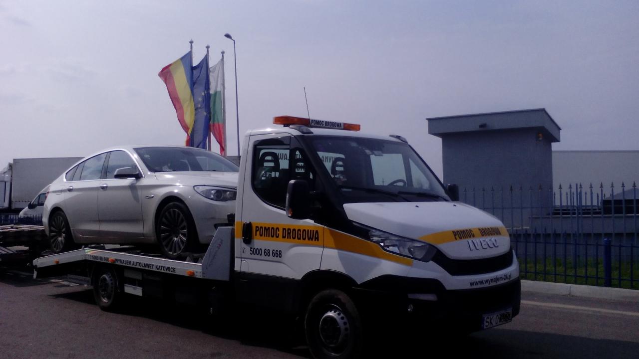 Laweta z białym BMW na platformie, na tle flagi UE, Rumunii i Bułgarii. Pomoc drogowa Iveco z napisem 'Pomoc Drogowa' i numerem telefonu.