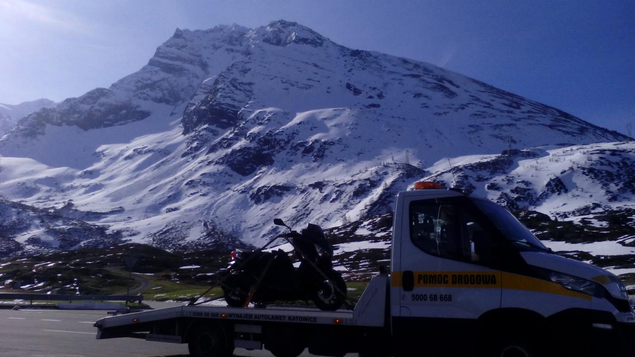 Laweta z motocyklem na tle ośnieżonych gór. Biała laweta z napisem 'Pomoc Drogowa' i numerem telefonu. Motocykl zabezpieczony pasami. Słoneczny dzień.