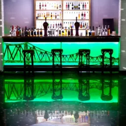 Podświetlony na zielono bar z motywem mostu, wysokie krzesła barowe, lustrzane odbicie w podłodze, półki z butelkami w tle.