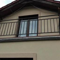 Czarna, metalowa balustrada balkonowa z pionowymi szczebelkami na balkonie domu jednorodzinnego z beżową elewacją i brązową stolarką okienną, widok z dołu.