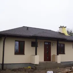 Gotowy parterowy dom z brązowym dachem i drewnianą stolarką okienną, widoczny komin zabezpieczony folią, stos drewna obok