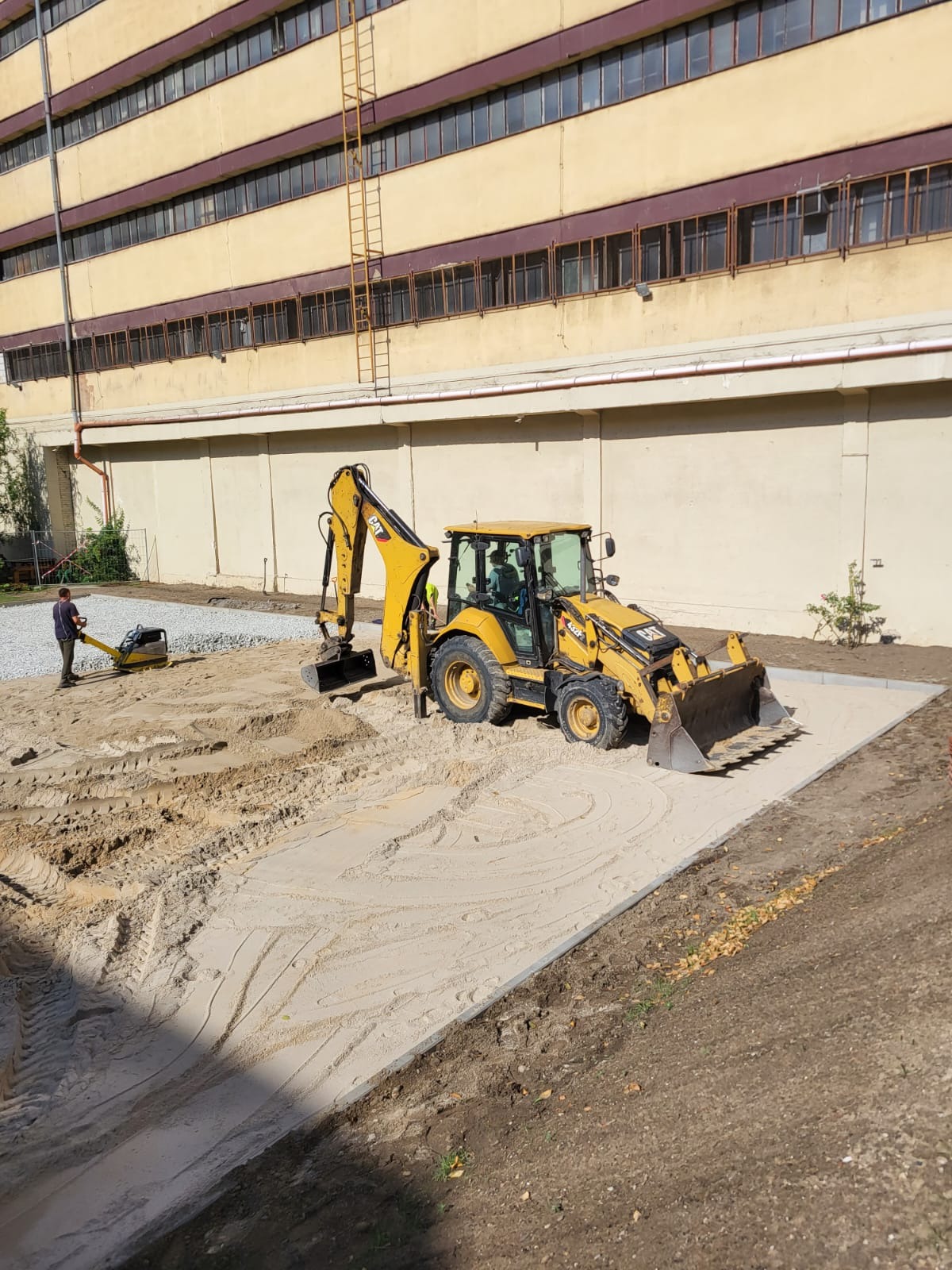 Żółta koparko-ładowarka CAT na placu budowy, gdzie pracownik zagęszcza podłoże pod przyszłe boisko sportowe na terenie Szpitala HCP w Poznaniu. W tle budynek szpitalny.