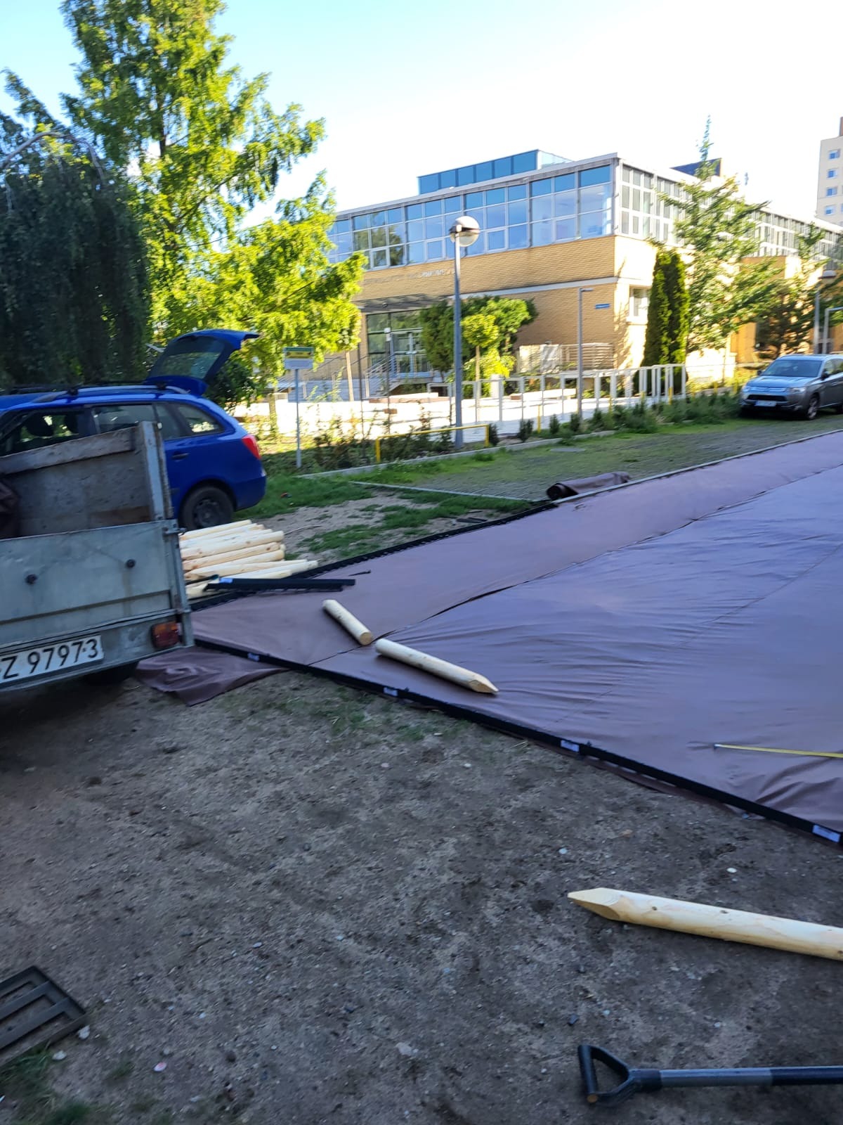 Przygotowanie podłoża pod trawnik z rolki: rozłożona ciemna folia na ziemi, drewniane kołki wbite wzdłuż krawędzi, przyczepka samochodowa z ładunkiem drewna w tle budynku.