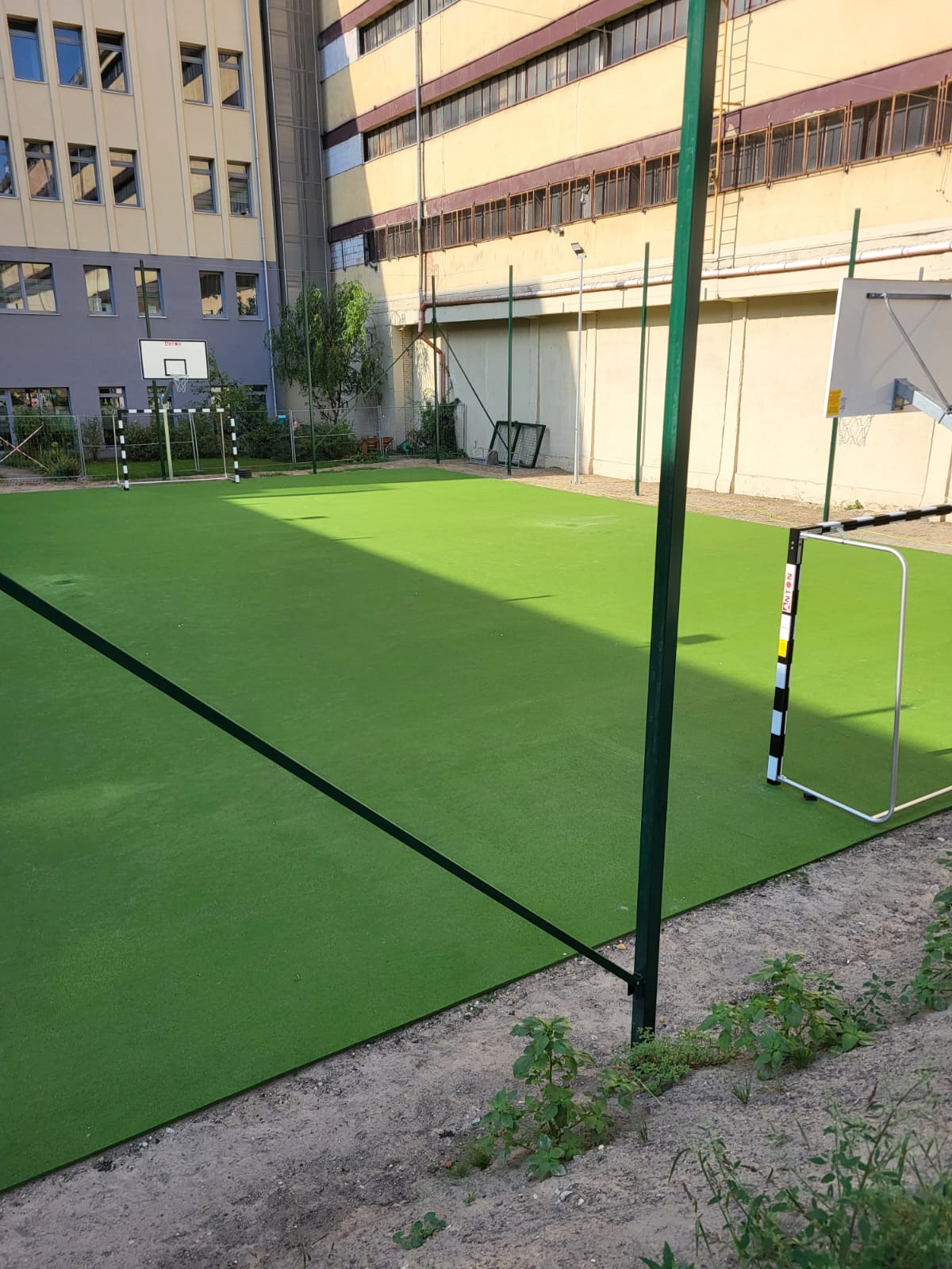 Boisko sportowe z zieloną sztuczną nawierzchnią, bramkami do piłki nożnej i koszem do koszykówki na tle budynku.