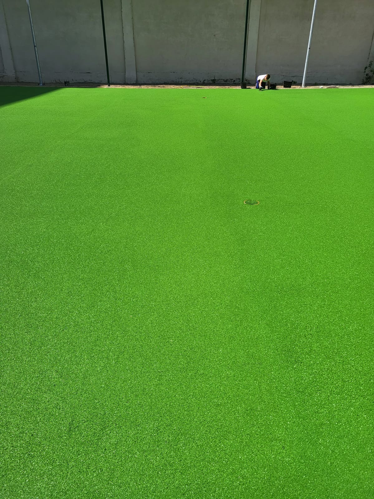 Świeżo ułożona, intensywnie zielona, sztuczna trawa na dużej powierzchni, z widoczną osobą pracującą przy krawędzi i dwoma okrągłymi elementami na trawie.
