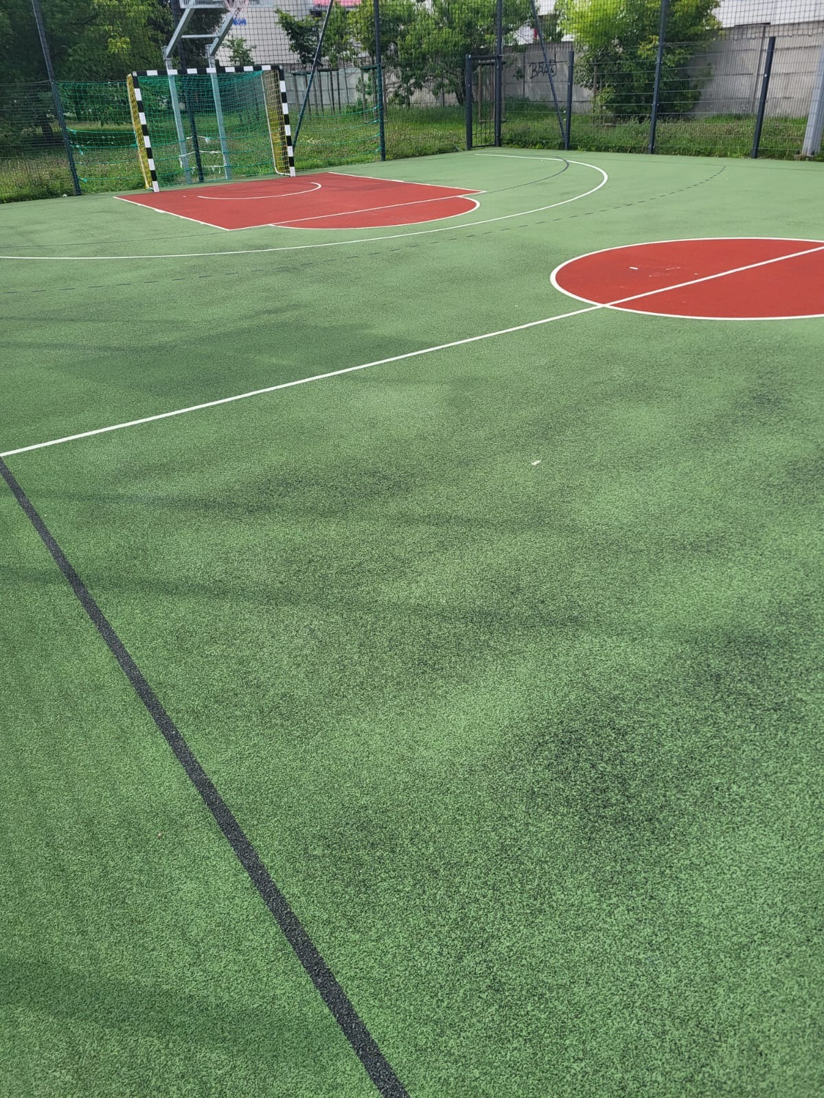 Zielone boisko sportowe z syntetycznej nawierzchni z elementami czerwonymi, widoczna bramka do piłki ręcznej, otoczone siatką.