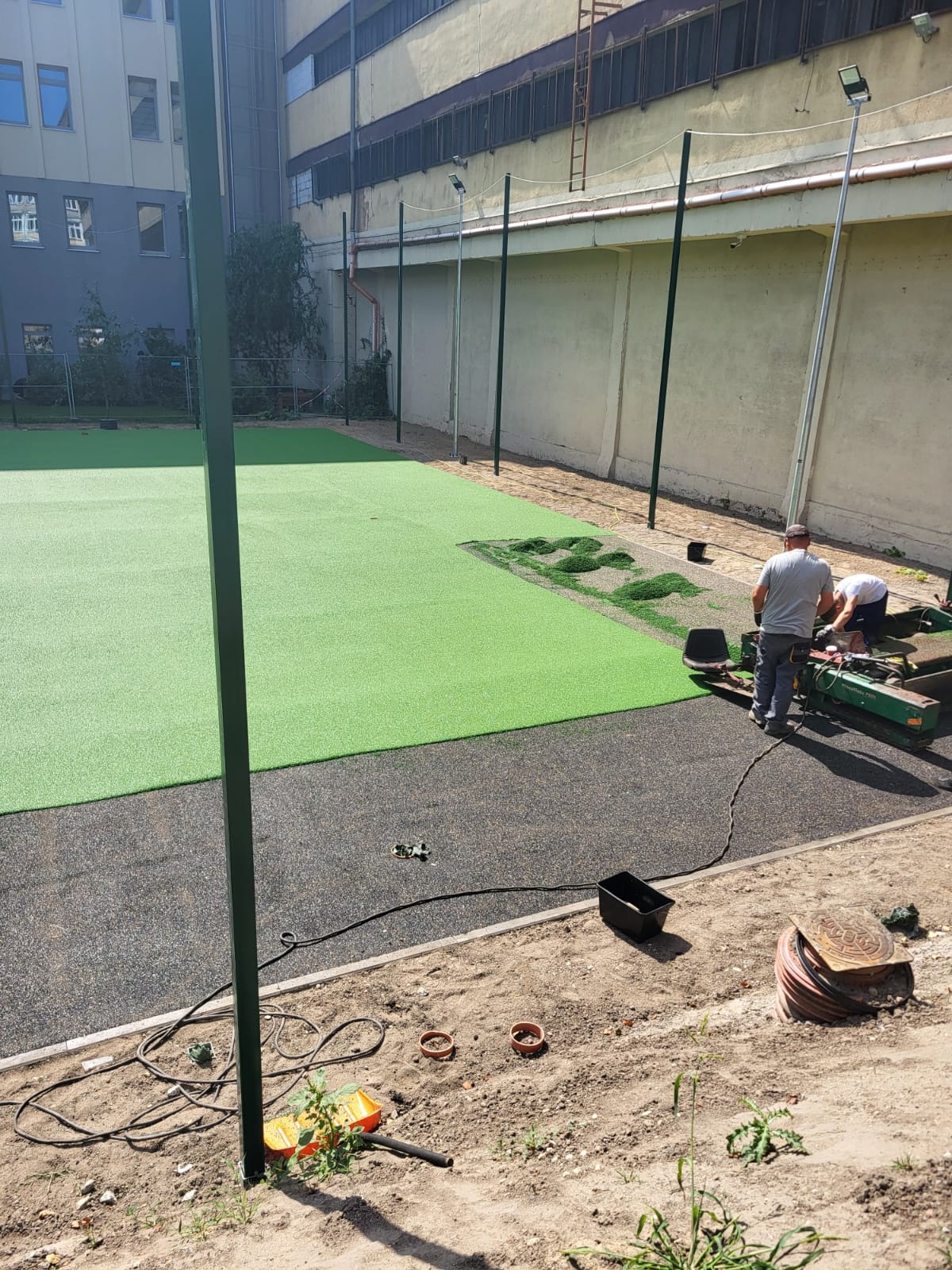 Montaż zielonej nawierzchni syntetycznej na boisku sportowym, widoczne narzędzia i osoby pracujące przy łączeniu fragmentów trawy, w tle budynek.