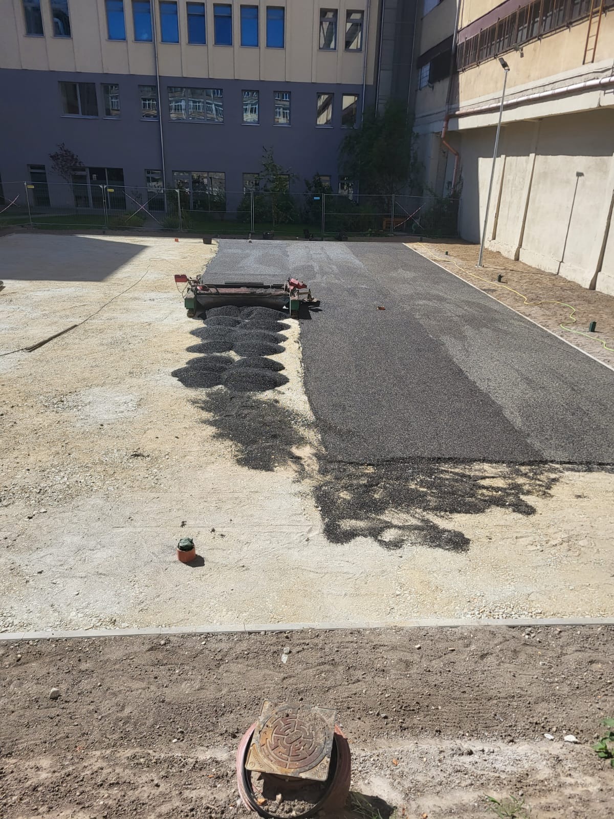 Rozkładanie asfaltu na placu za pomocą maszyny, widoczne nierówności i grudki asfaltu, w tle budynek.