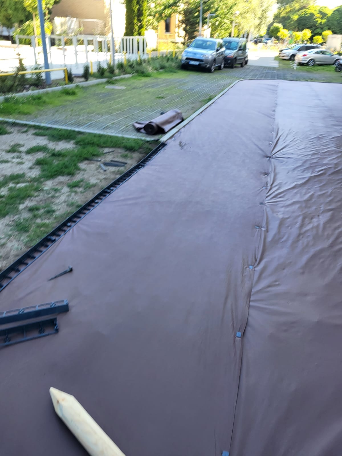 Rozkładanie brązowej trawy z rolki na przygotowanym podłożu z obrzeżami, widoczne elementy mocujące i częściowo zwinięta rolka w tle.