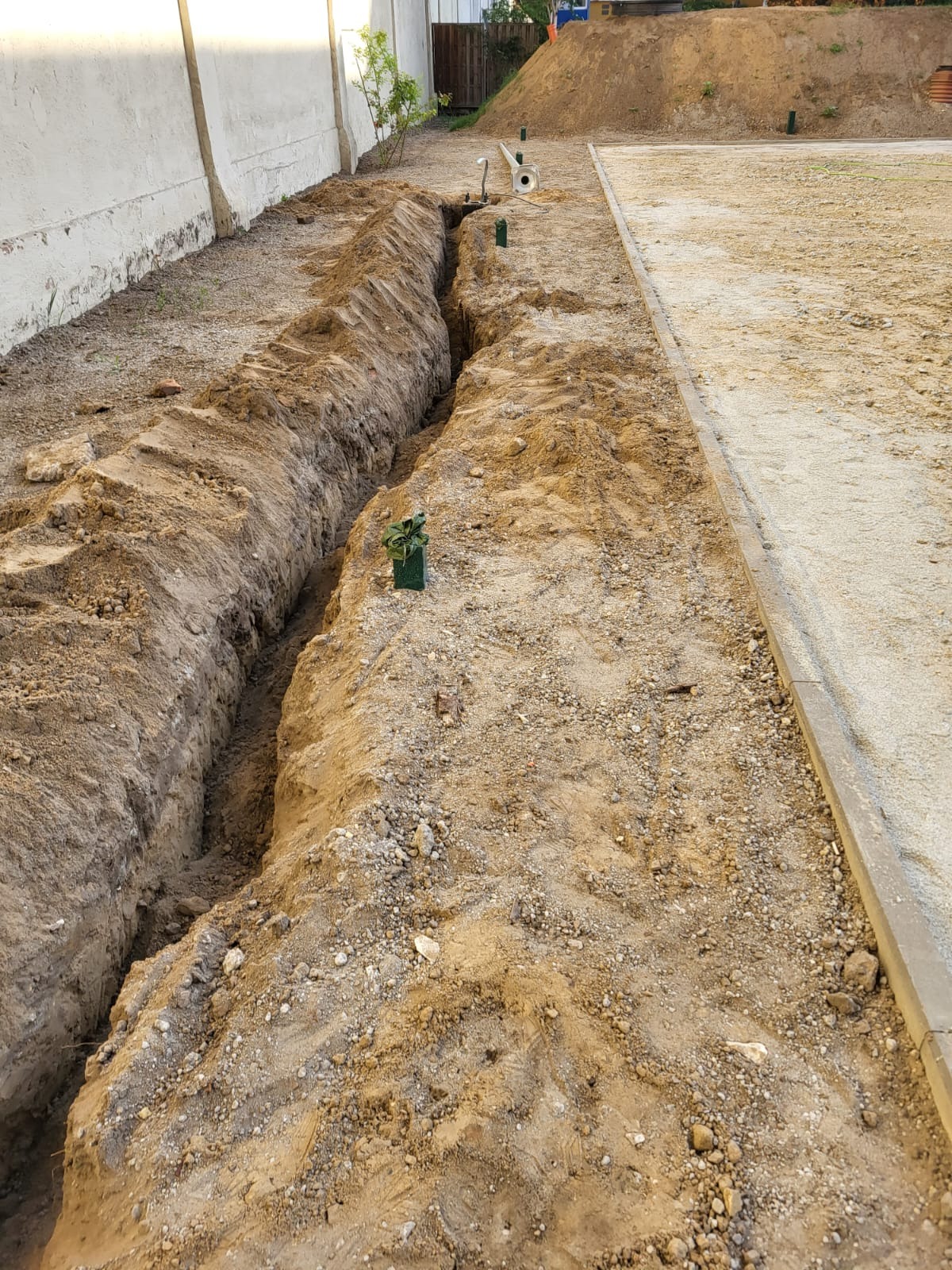 Wykopany rów w ziemi przygotowany pod instalację w ogrodzie, widoczny fragment trawnika z obrzeżem oraz roślina w doniczce przy krawędzi rowu.