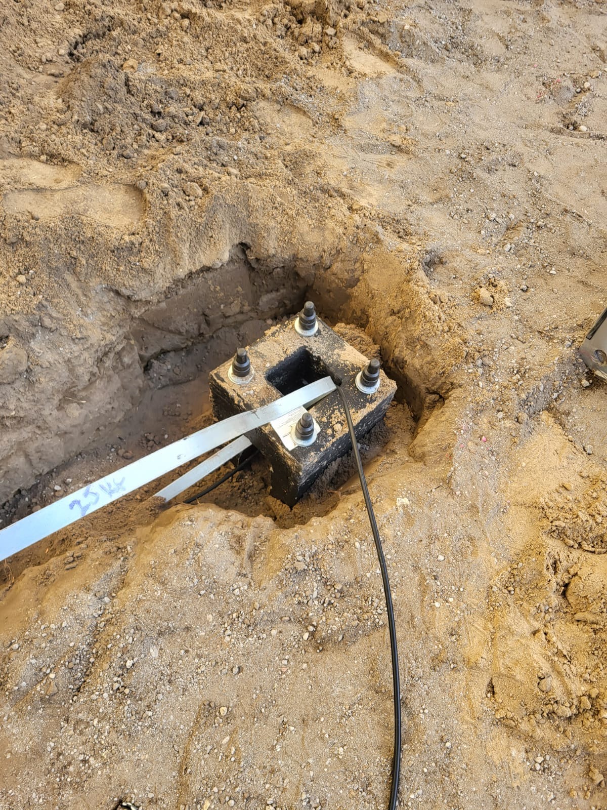 Przygotowany fundament pod plac zabaw z metalowymi mocowaniami i przewodami elektrycznymi, umieszczony w wykopie wypełnionym piaskiem.