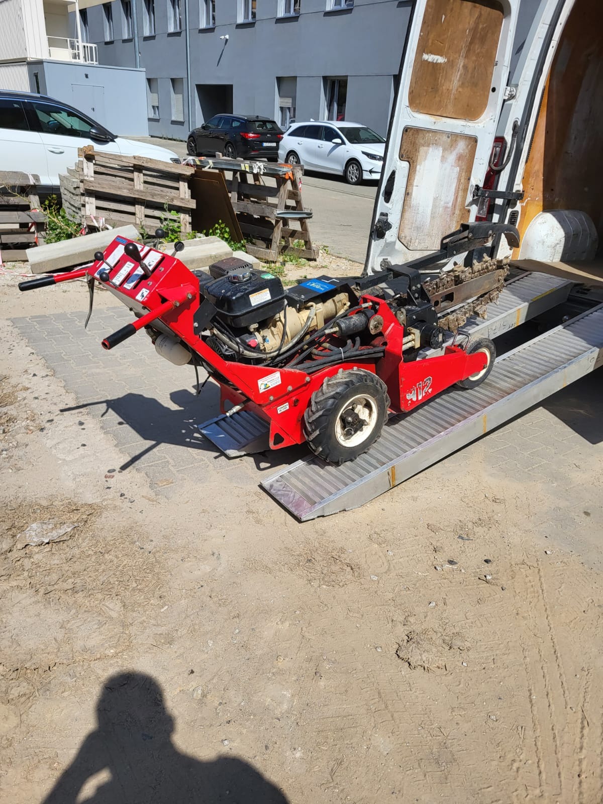 Czerwona glebogryzarka łańcuchowa na rampie wewnątrz furgonetki, gotowa do transportu, z widocznymi elementami sterowania i silnikiem spalinowym, na tle miejskiej zabudowy.