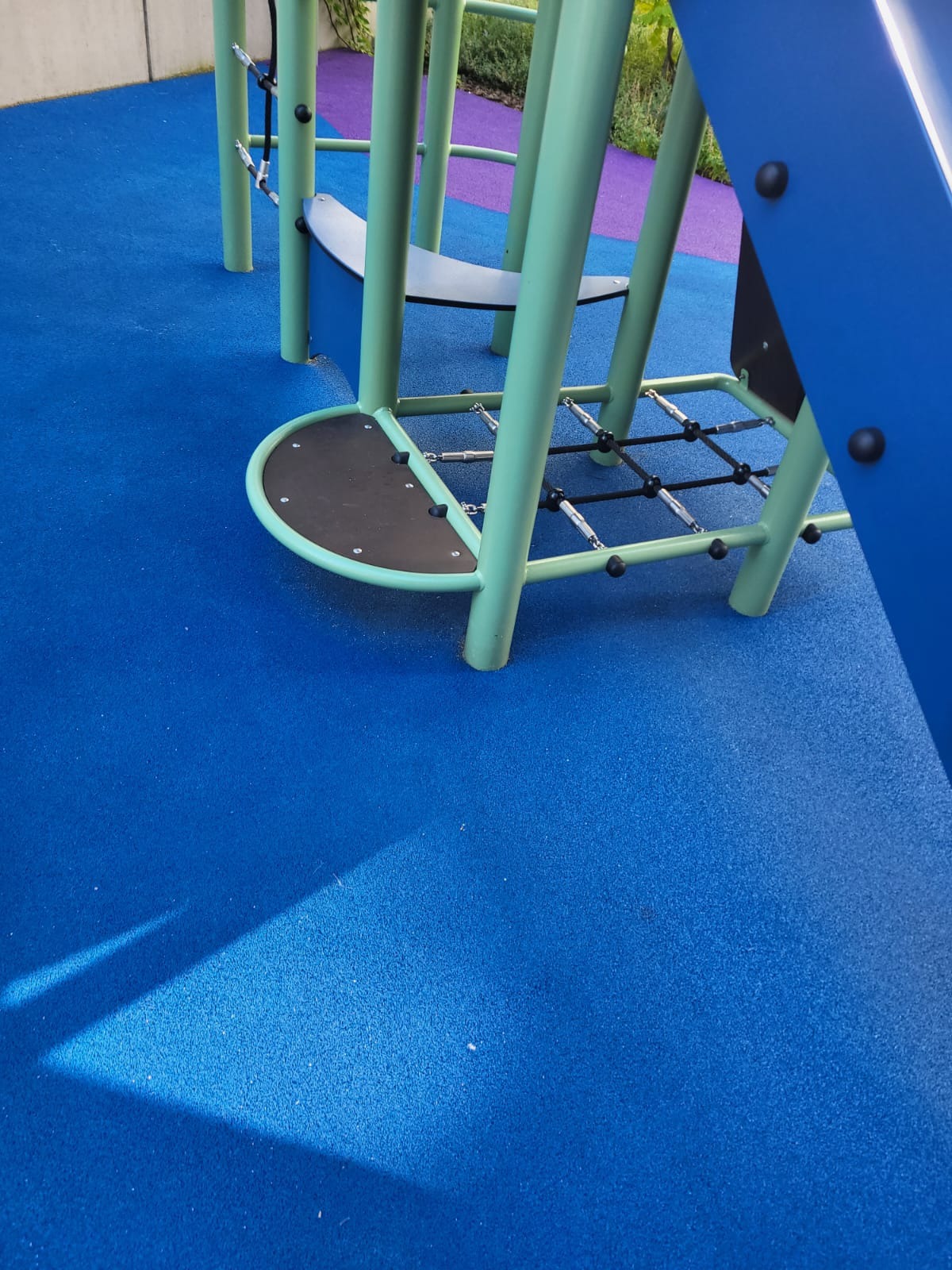 Fragment konstrukcji placu zabaw z metalowymi rurkami w kolorze seledynowym, czarnymi elementami i niebieską, gumową nawierzchnią amortyzującą upadki.