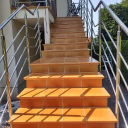 Zewnętrzne schody wejściowe wyłożone pomarańczowymi płytkami ceramicznymi z błyszczącą, metalową balustradą, prowadzące do drzwi wejściowych domu.