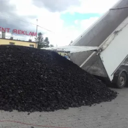 Wyładunek węgla z naczepy ciężarówki na wybrukowany plac, widoczny budynek z napisem 'PRODUCENT REKLAM' w tle.