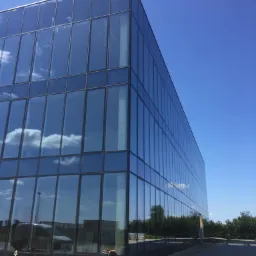 Nowoczesny budynek biurowy z fasadą ze szkła refleksyjnego, odbijającego błękitne niebo z chmurami, widoczne prace wykończeniowe na zewnątrz.