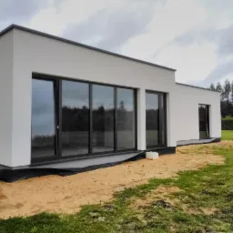 Nowoczesny biały dom z dużymi przesuwnymi oknami z ciemnymi ramami, widok z zewnątrz na trawnik i las w tle, widoczne prace ziemne przy fundamentach.