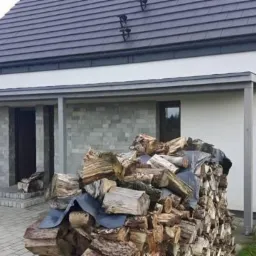 Nowoczesny dom z szarą pergolą przy wejściu, obok stos drewna opałowego przykryty folią.