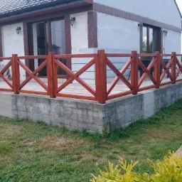 Zewnętrzny taras z drewnianą balustradą w kształcie krzyży, osadzony na betonowej podstawie, przylegający do domu z brązowymi i białymi elementami elewacji.