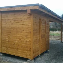 Drewniana altana z czarnym dachem i rynnami, postawiona na betonowych bloczkach, widok z boku na tle żwirowego podłoża i fragmentu budynku.