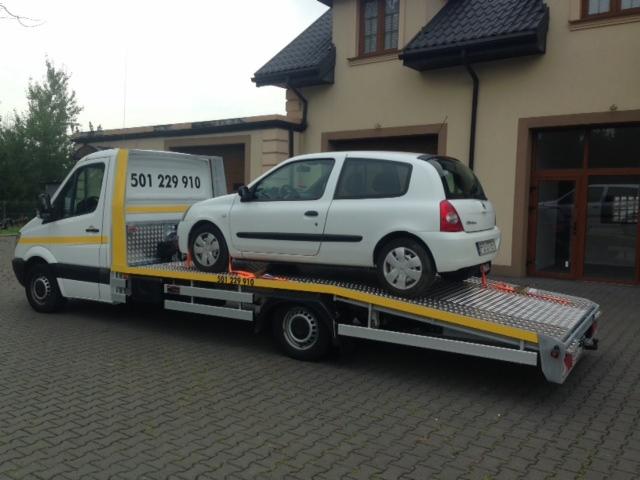 Biały samochód osobowy Renault Clio zabezpieczony pomarańczowymi pasami transportowymi na platformie lawety z numerem telefonu 501 229 910 na zabudowie, na tle budynku z brązową stolarką okienną.