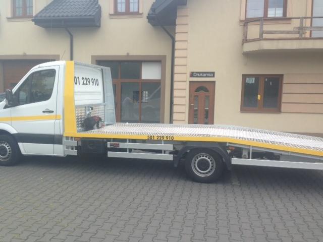 Biała laweta marki Mercedes z żółtymi elementami karoserii zaparkowana na brukowanej ulicy przed budynkiem z napisem 'Drukarnia' nad drzwiami.