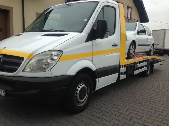 Biała laweta Mercedes z żółtymi pasami transportuje biały samochód osobowy na tle brukowanej drogi i budynku.