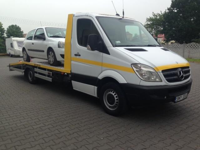 Biała laweta Mercedes z żółtymi pasami transportuje biały samochód osobowy Renault Clio na tle brukowanej powierzchni i zieleńców.