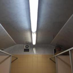 Zano - Wnętrze kontenera sanitarnego z widoczną instalacją elektryczną, wentylacją i częściowo wykończonymi ścianami z płytek ceramicznych. Widoczny fragment sufitu z paneli i oświetlenie jarzeniowe.