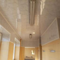 Zano - Błyszczący sufit z paneli imitujących marmur w korytarzu z żółtymi płytkami ściennymi, widoczne lampy fluorescencyjne i wieszaki na ścianie.