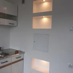 Biała ściana z wnękami podświetlonymi punktowymi lampami LED, obok fragmentu zabudowy kuchennej z białymi frontami i metalowym zlewem.