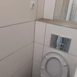 Wykończona łazienka w Przystajni z jasnymi płytkami ceramicznymi, widoczny fragment toalety i przycisk spłuczki.