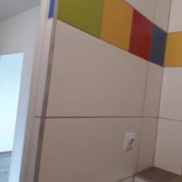 Zano - Fragment ściany z płytami wykończeniowymi w odcieniach beżu, z poziomymi fugami, u góry dekoracyjny pas z kolorowych kwadratów (zielony, żółty, czerwony, niebieski), widoczne gniazdko elektryczne...