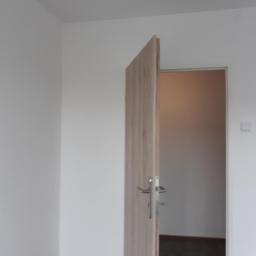 Zano - Świeżo pomalowane, białe ściany i częściowo otwarte drzwi z klamką w jasnym odcieniu drewna, ukazujące fragment podłogi w pomieszczeniu obok.