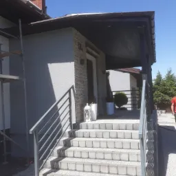 Wykończone schody wejściowe do domu w Przystajni, z szarą elewacją i dekoracyjnym kamieniem, widoczne rusztowanie sugeruje prace wykończeniowe.