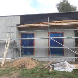 Zano - Surowa elewacja budynku w trakcie prac tynkarskich, okna zabezpieczone folią, widoczne rusztowania i materiały budowlane na trawniku.