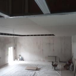 Zano - Wnętrze w trakcie montażu sufitu podwieszanego z płyty karton-gips, widoczne profile metalowe i częściowo zamontowane płyty, pomieszczenie zabezpieczone folią malarską.