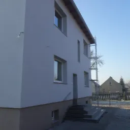 Dwupiętrowy dom po ociepleniu elewacji, z widocznymi schodami wejściowymi z szarego kamienia i częściowo rozstawionym rusztowaniem z tyłu budynku.