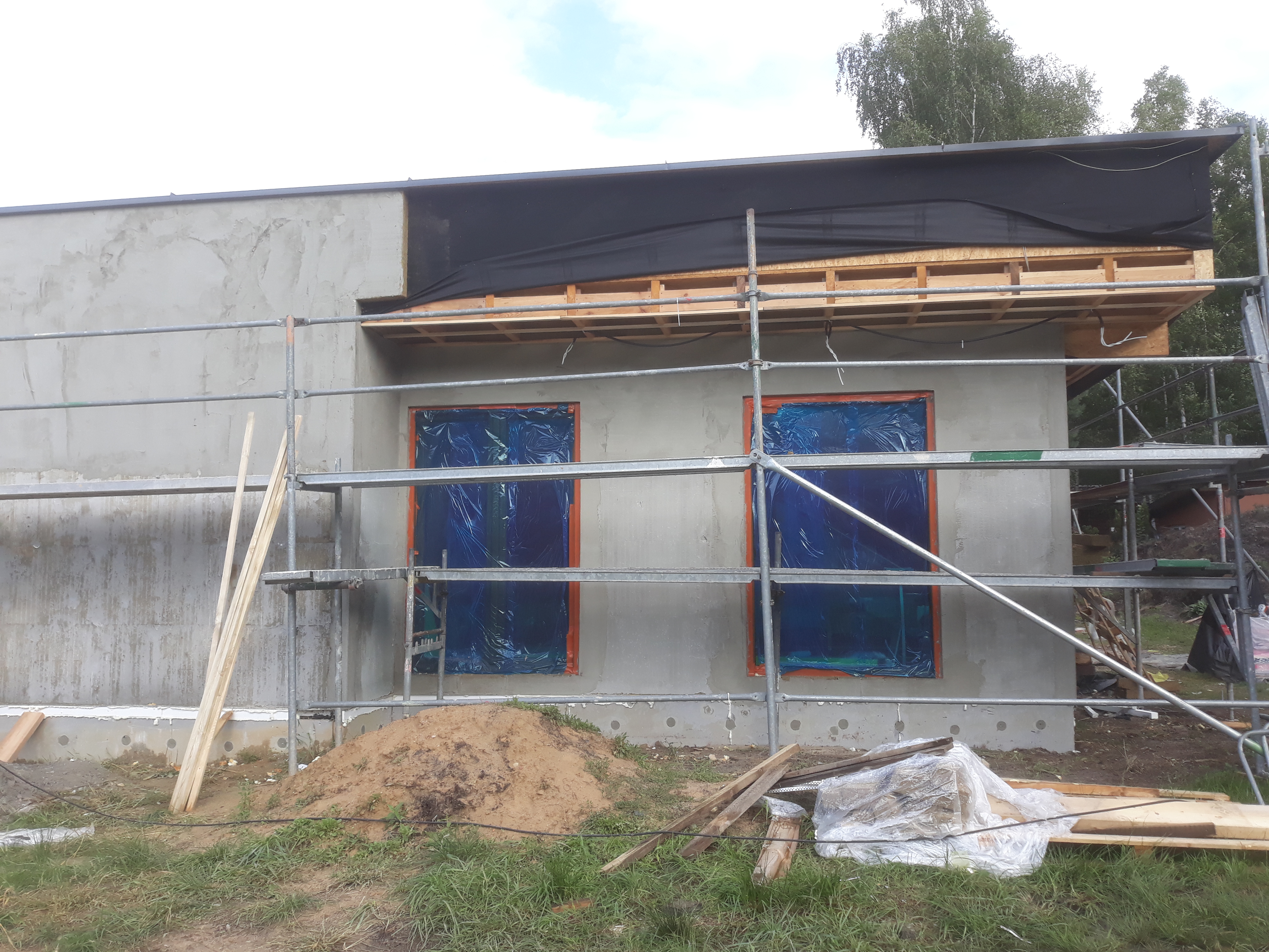 Surowa elewacja budynku w trakcie prac tynkarskich, okna zabezpieczone folią, widoczne rusztowania i materiały budowlane na trawniku.