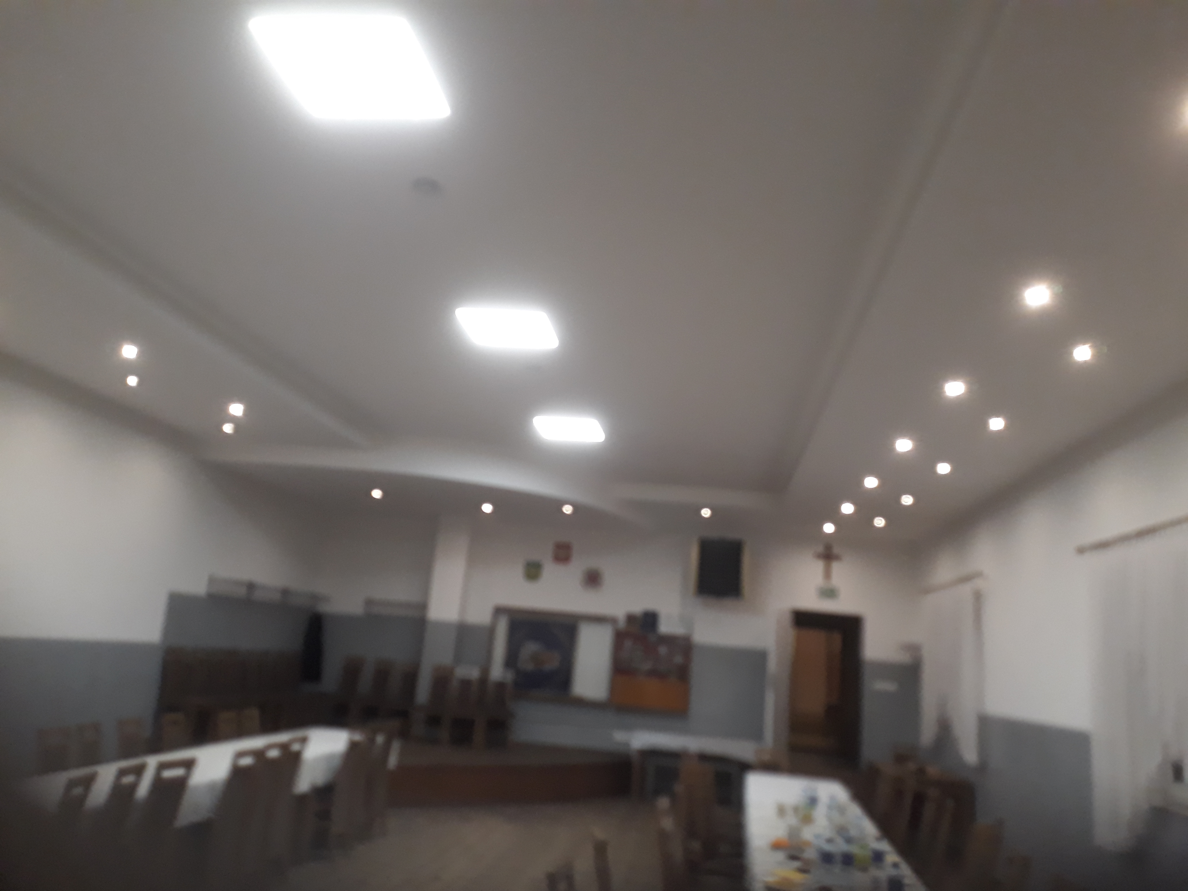 Wnętrze sali z białym sufitem, oświetleniem punktowym i kwadratowymi lampami, widoczne stoły z krzesłami i dekoracje na ścianach.