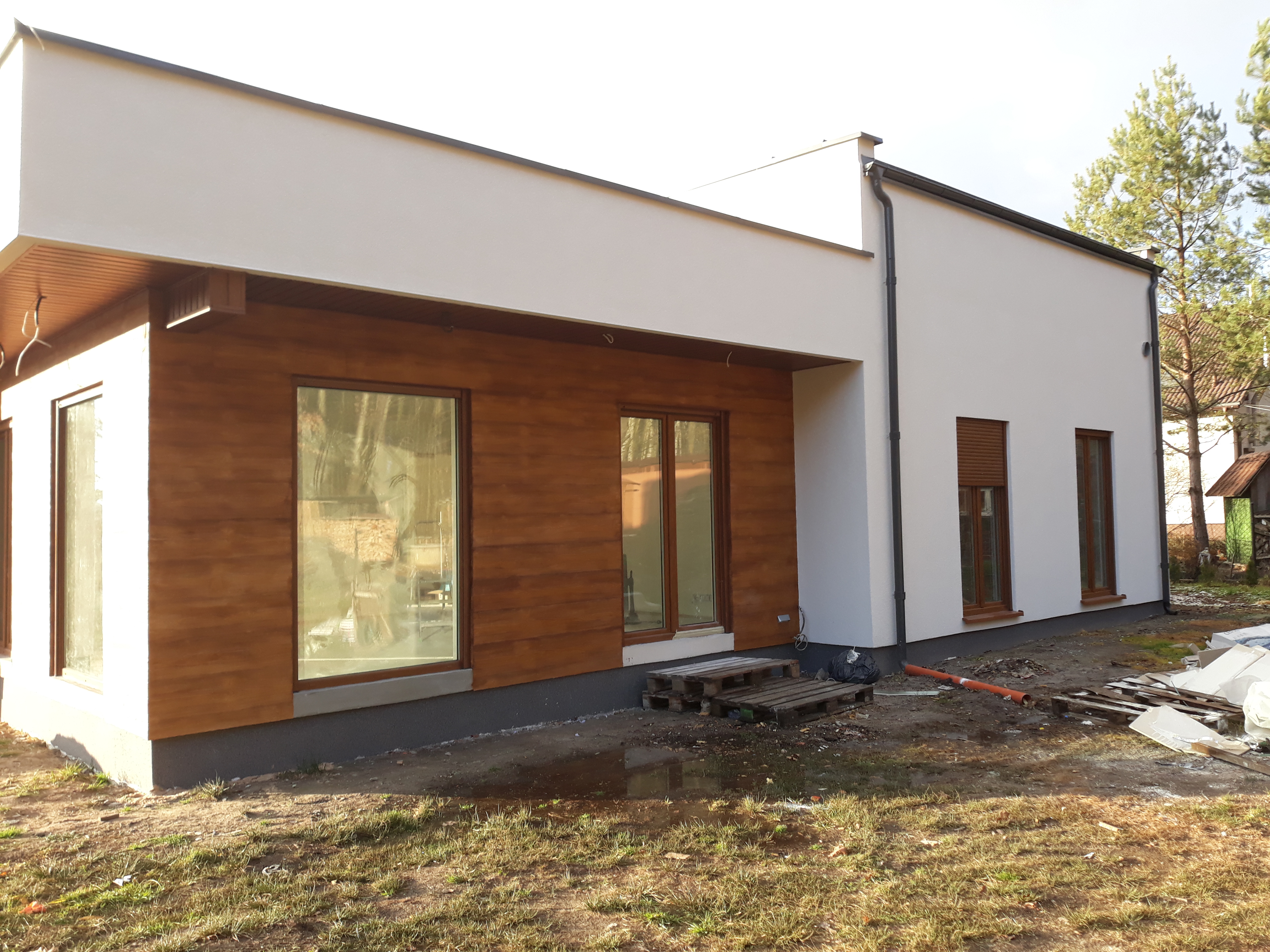 Nowoczesny dom jednorodzinny z elewacją częściowo wykończoną drewnopodobnymi panelami, białe ściany, brązowe okna, teren wokół w trakcie prac wykończeniowych, widoczne palety i materiały budowlane.