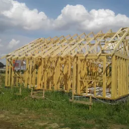 Surowa konstrukcja drewniana domu w trakcie budowy, widoczne fundamenty i szkielet ścian oraz dachu, na pierwszym planie trawa, w tle błękitne niebo z chmurami.