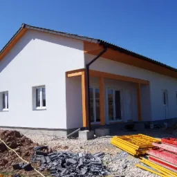 Niedawno otynkowany, parterowy budynek z drewnianą konstrukcją werandy i stertą dachówek oraz rusztowań w pobliżu, na tle błękitnego nieba.