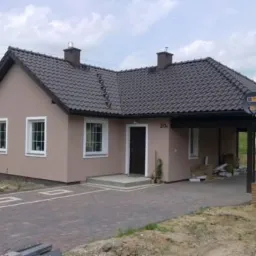 Niski, parterowy dom z beżową elewacją, ciemną dachówką i przyległą wiatą garażową. Widoczny fragment brukowanego podjazdu i trawnika. Na wiacie zamontowana antena satelitarna.