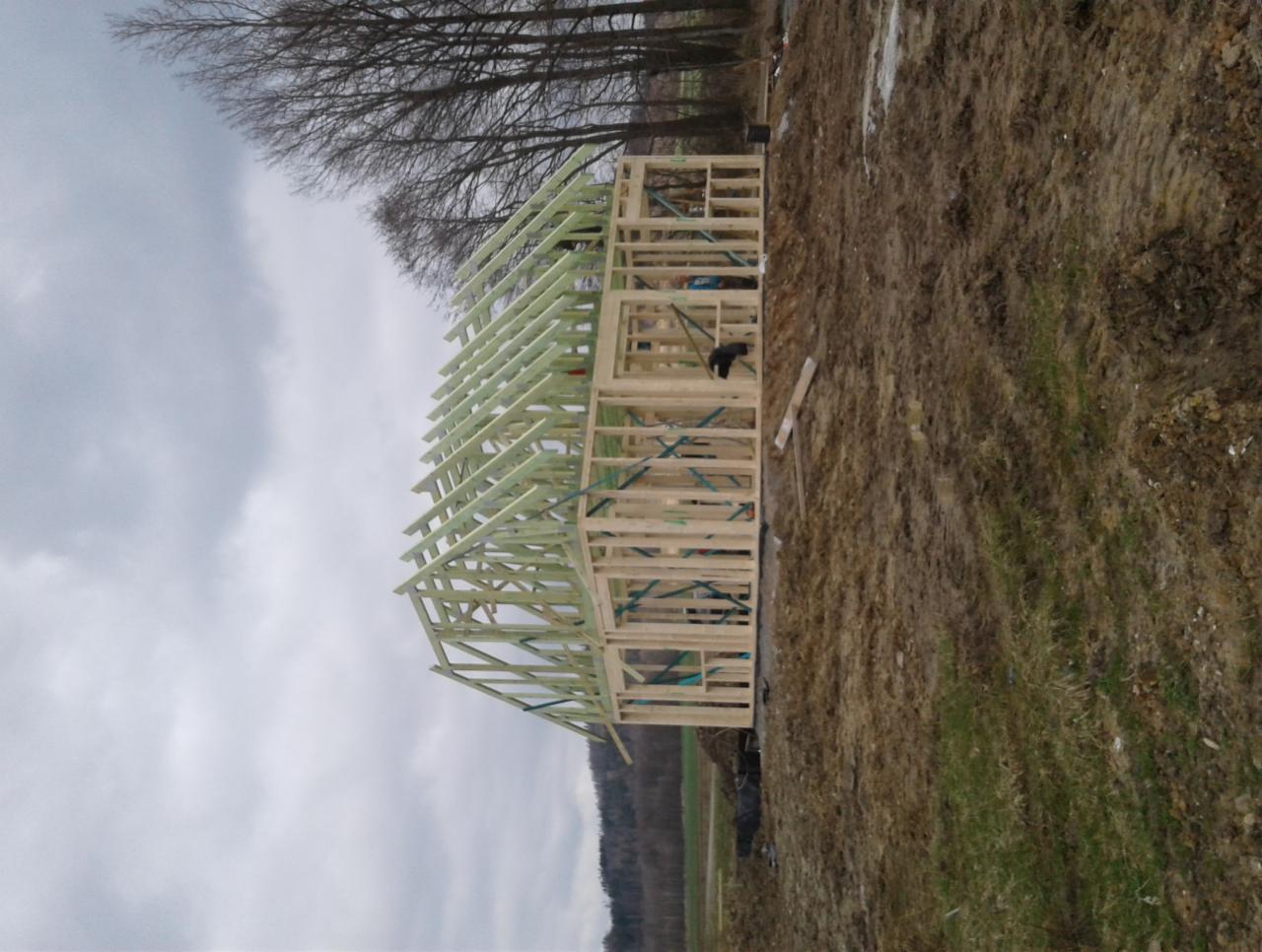 Wznoszenie szkieletu drewnianego domu z widocznymi elementami konstrukcyjnymi, teren budowy z błotnistą ziemią i zachmurzonym niebem w tle.