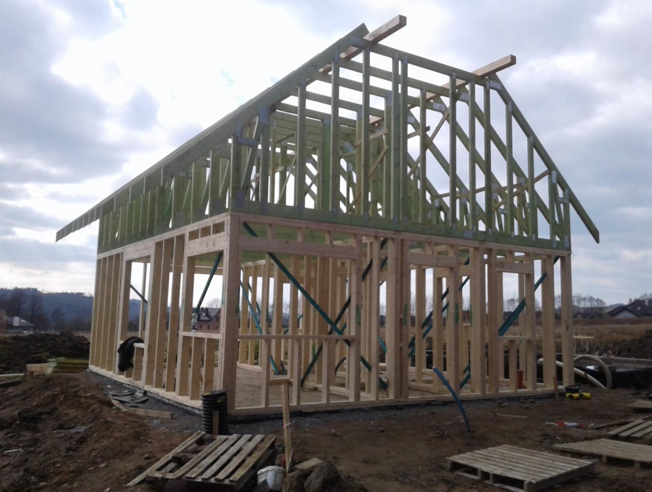 Konstrukcja szkieletowa domu w trakcie budowy, widoczne drewniane elementy ścian i dachu, zabezpieczone zielonym impregnatem, na tle pochmurnego nieba.
