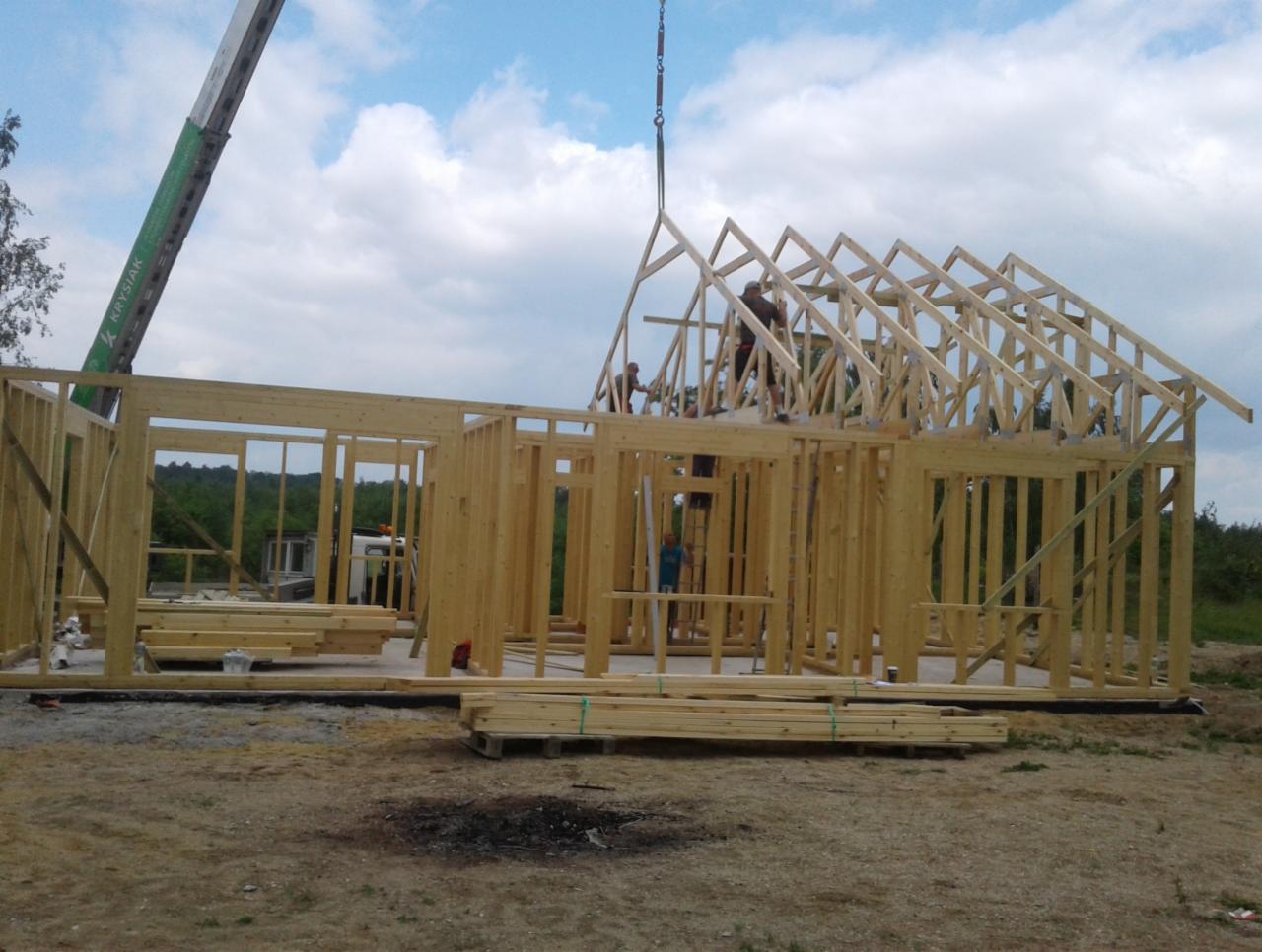 Montaż więźby dachowej na konstrukcji budynku szkieletowego z użyciem dźwigu, widoczni pracownicy na dachu i elementy konstrukcyjne na ziemi.