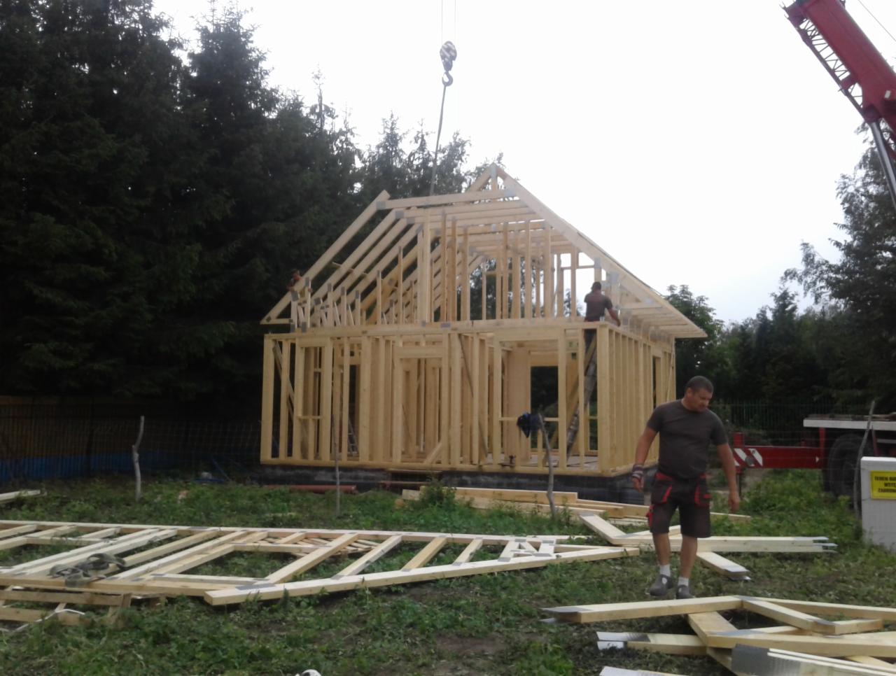 Wznoszenie szkieletu drewnianego domu jednorodzinnego z użyciem dźwigu, widoczny pracownik na drabinie oraz elementy konstrukcyjne na trawie.