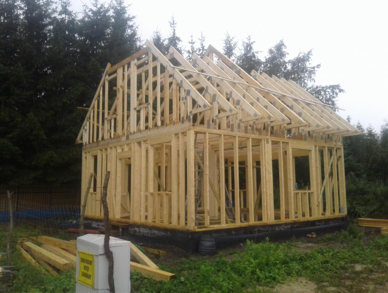 Surowa konstrukcja domu z drewna na fundamencie, widoczne elementy szkieletu ścian i dachu, w tle las iglasty i tablica ostrzegawcza.