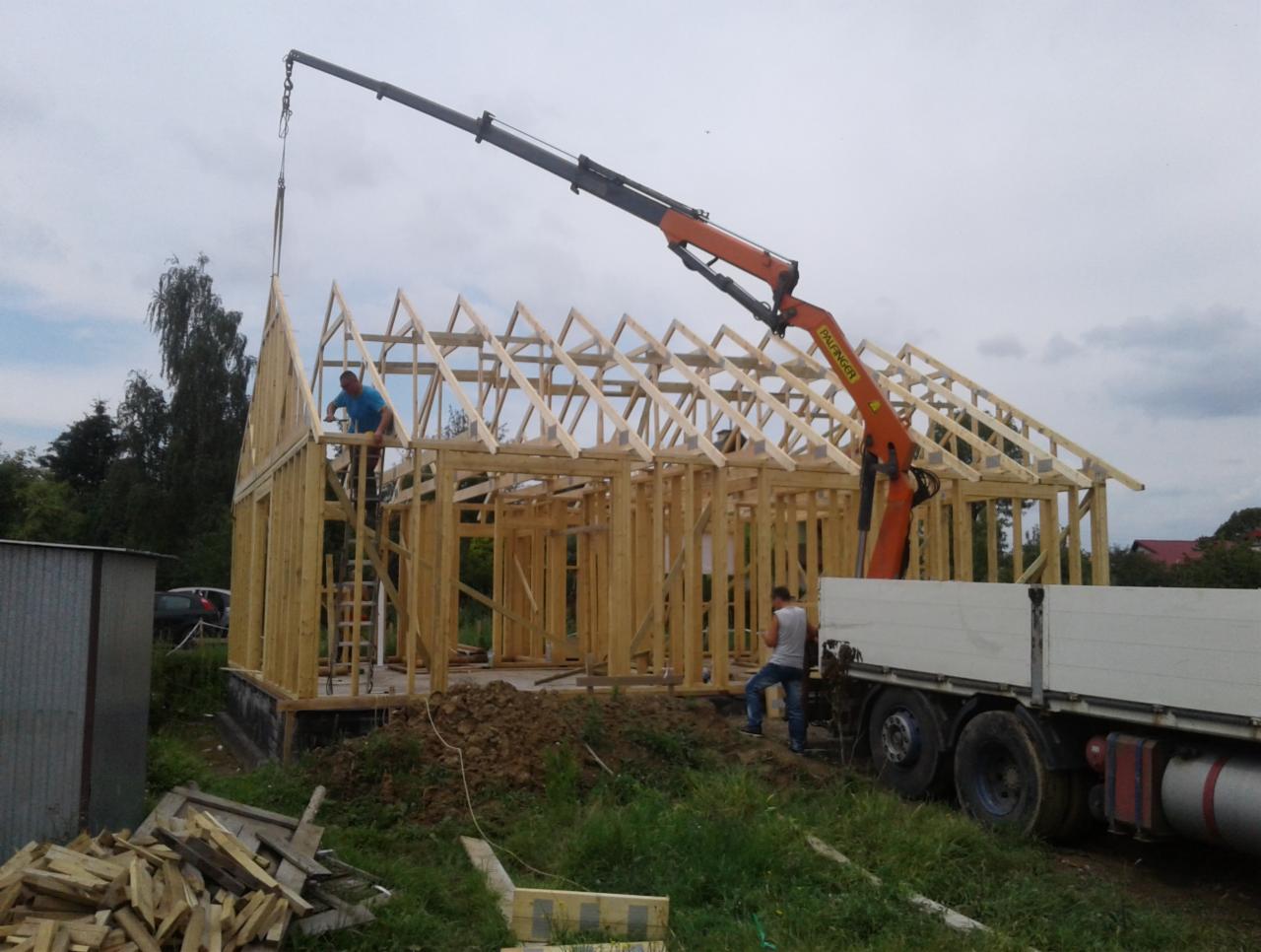 Montaż konstrukcji szkieletowej domu jednorodzinnego z użyciem dźwigu Palfinger, widoczne elementy więźby dachowej i pracownicy na budowie.